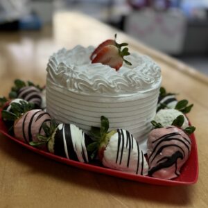Small white Valentine’s cake topped with a strawberry, served on a red heart shaped plate surrounded by assorted chocolate covered strawberries with pink, white, and dark chocolate drizzle.