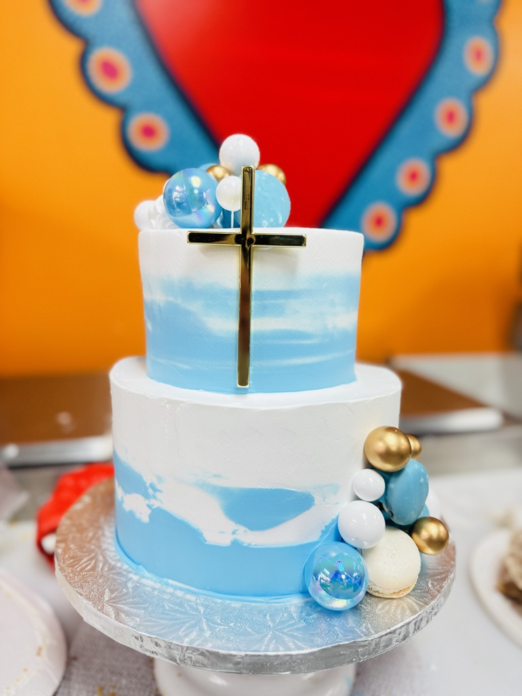 Two-tier baptism cake with white and sky-blue buttercream, a gold cross on the top tier, and blue, white, and gold balloon-style decorations with a white macaron on the side.
