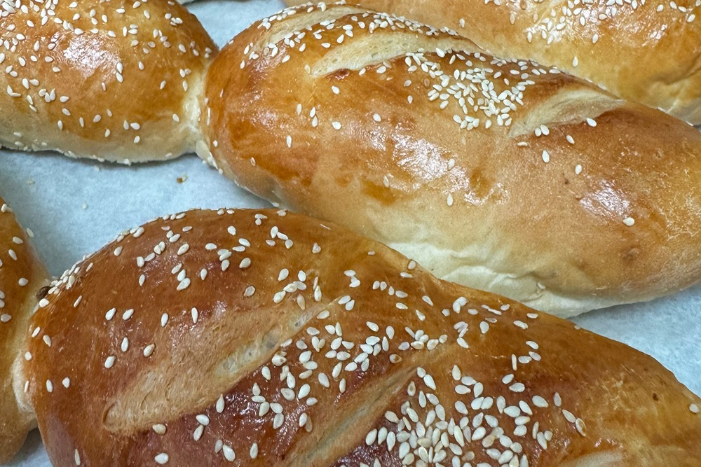 Freshly baked bolillo rolls with a golden crust, diagonal scoring, and sesame seeds on a baking tray.