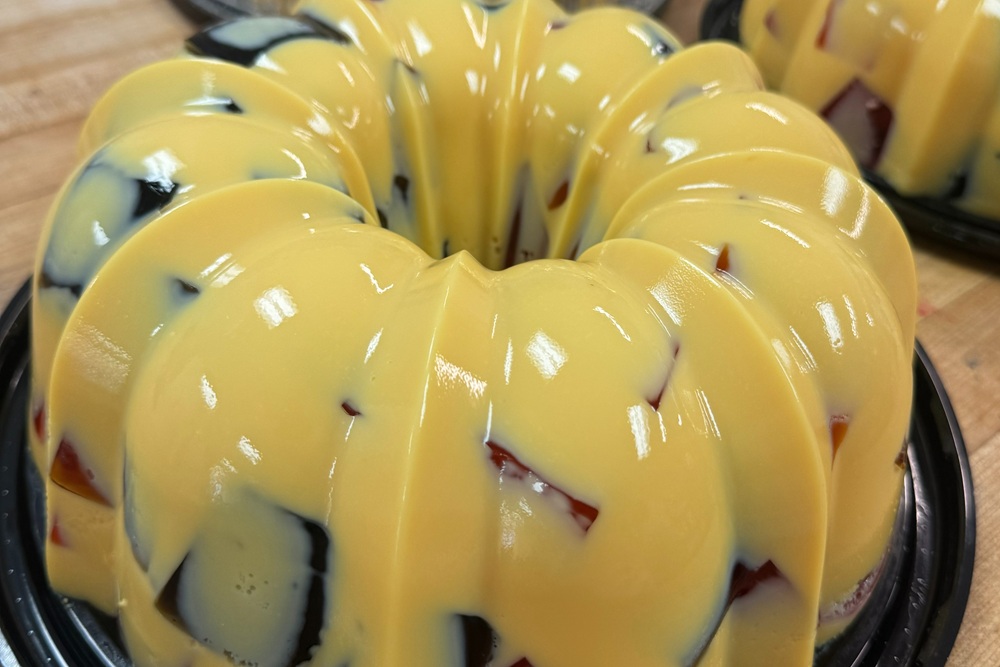 Glossy ring-shaped gelatin dessert with a creamy yellow base and colorful fruit pieces, set in a bundt-style mold on a black tray.