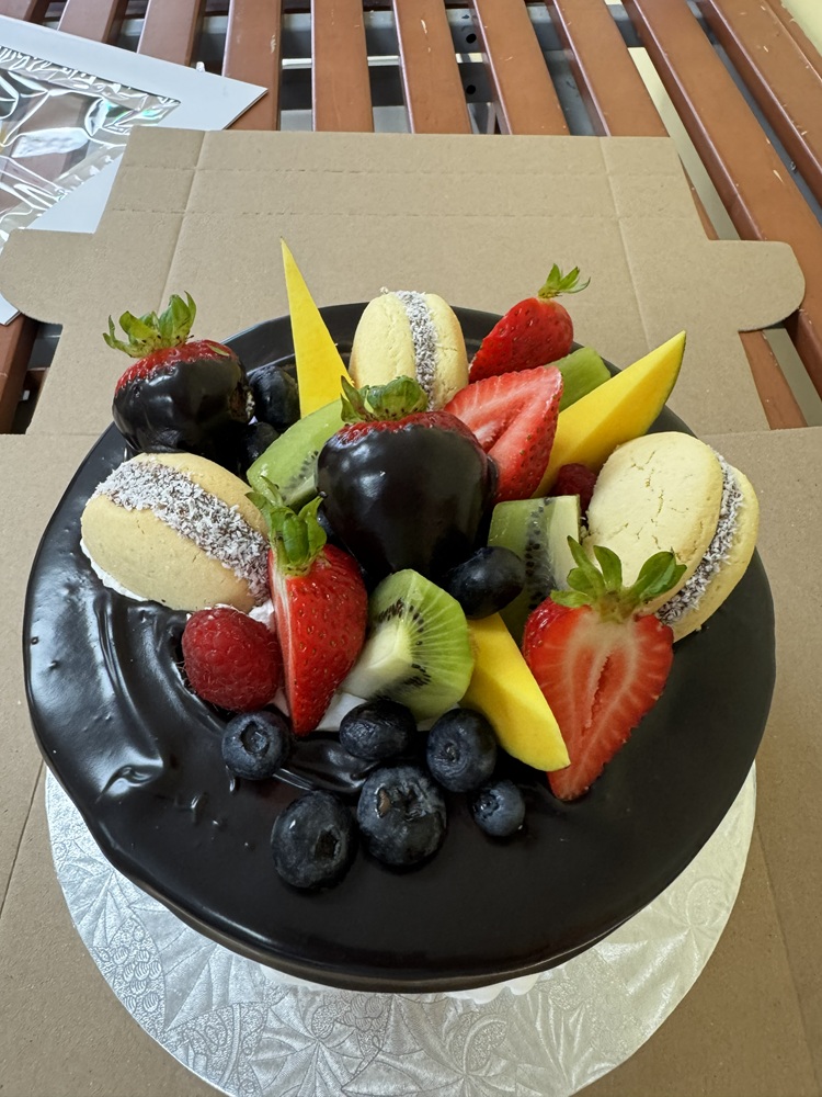 Chocolate glazed cake topped with fresh strawberries, blueberries, kiwi, mango slices, raspberries, and coconut-covered cookies, packaged in a bakery box.