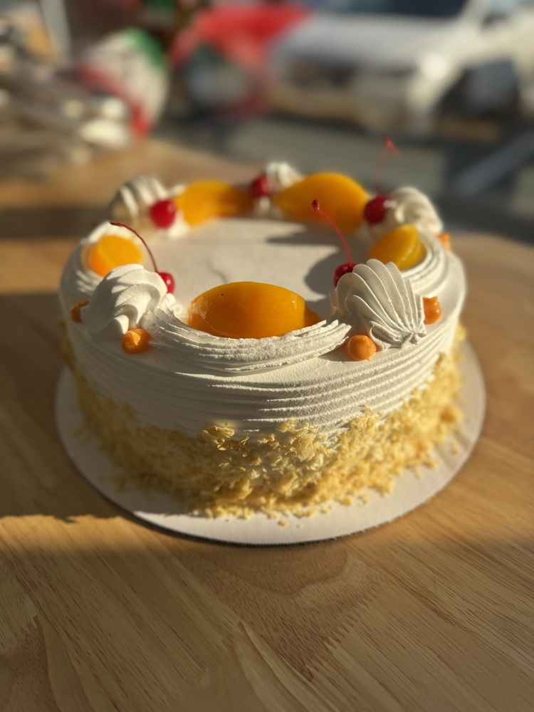 Round peach and cherry cake with white whipped frosting, peach slices and cherries on top, and crushed cake crumbs around the sides.