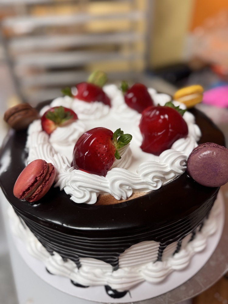 Chocolate drip cake with whipped cream swirls, glazed strawberries on top, and assorted macarons around the edge.