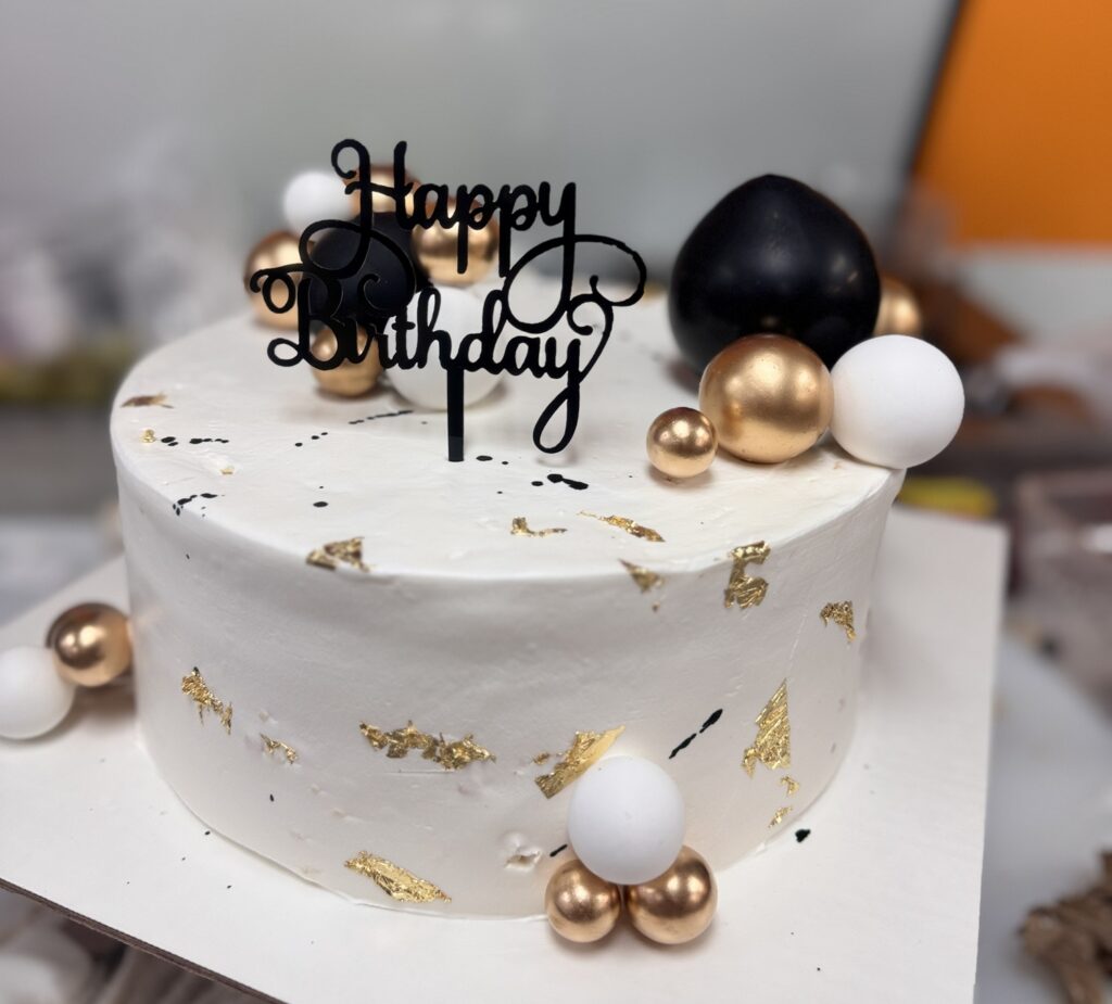 A white frosted birthday cake with gold leaf accents, a black “Happy Birthday” topper, and gold, white, and black decorative spheres.