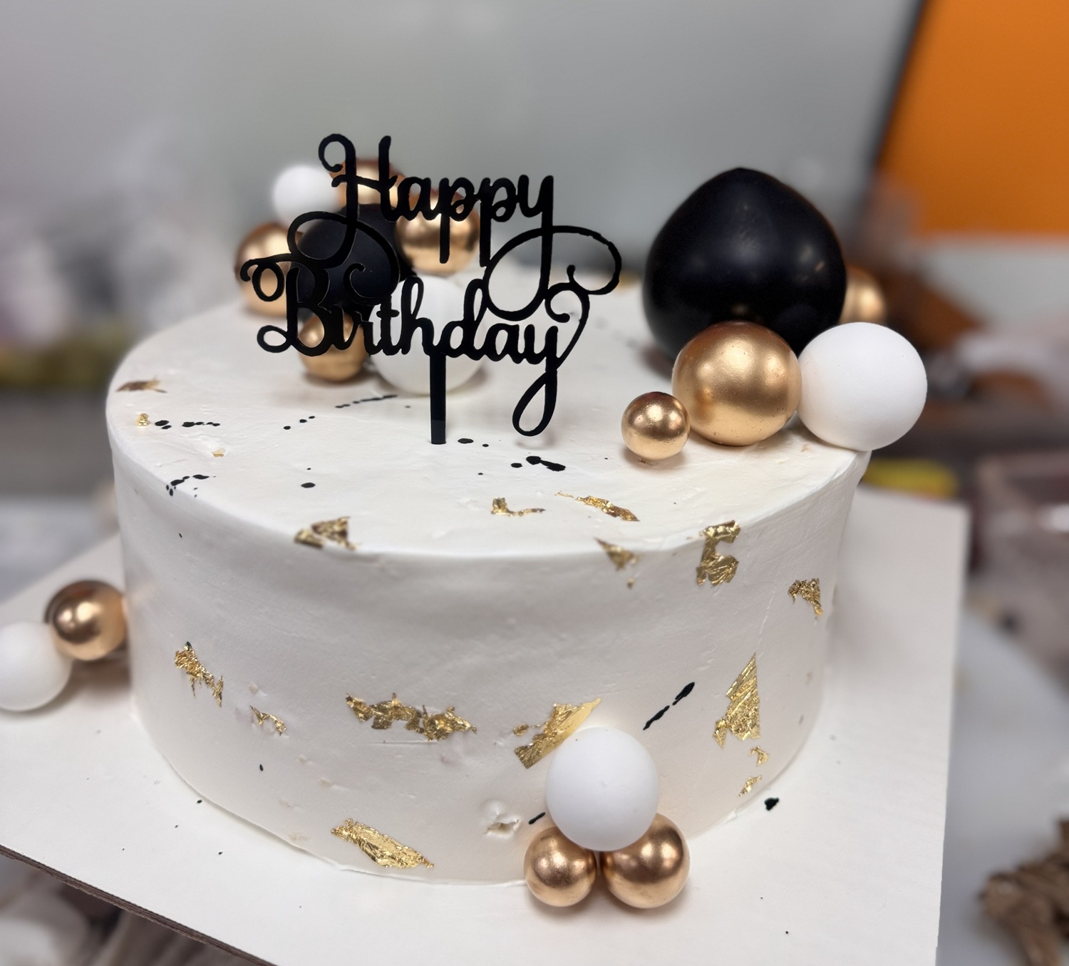 A white frosted birthday cake with gold leaf accents, a black “Happy Birthday” topper, and gold, white, and black decorative spheres.