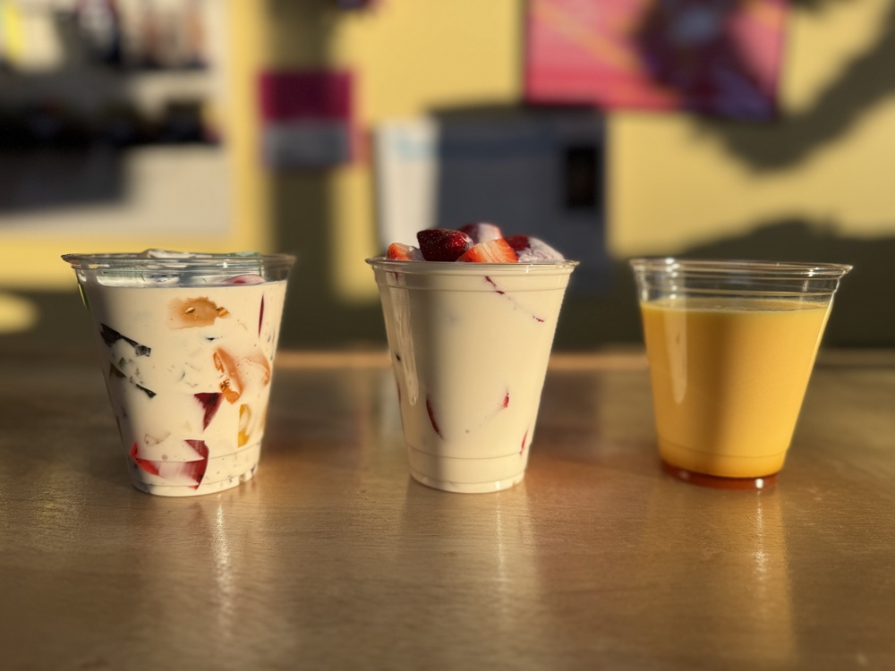 Three clear cups on a table, including a creamy jello salad with colorful gelatin pieces, a creamy fruit cup topped with strawberries, and a cup of yellow juice.