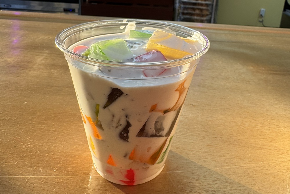 Clear plastic cup filled with creamy jello salad and colorful gelatin cubes, sitting on a wooden counter.