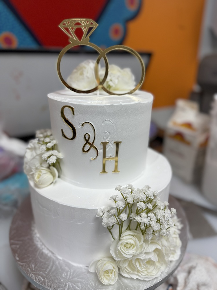 Two-tier white wedding cake with smooth buttercream, gold monogram letters “S & H” on the top tier, a gold interlocking rings topper, and white roses with baby’s breath arranged around the base.