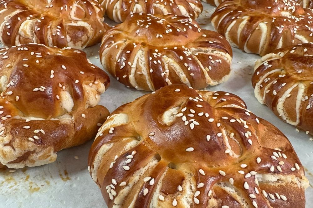 Freshly baked pan de feria breads with a glossy golden crust, decorative slits, and sesame seeds, cooling on a baking tray.